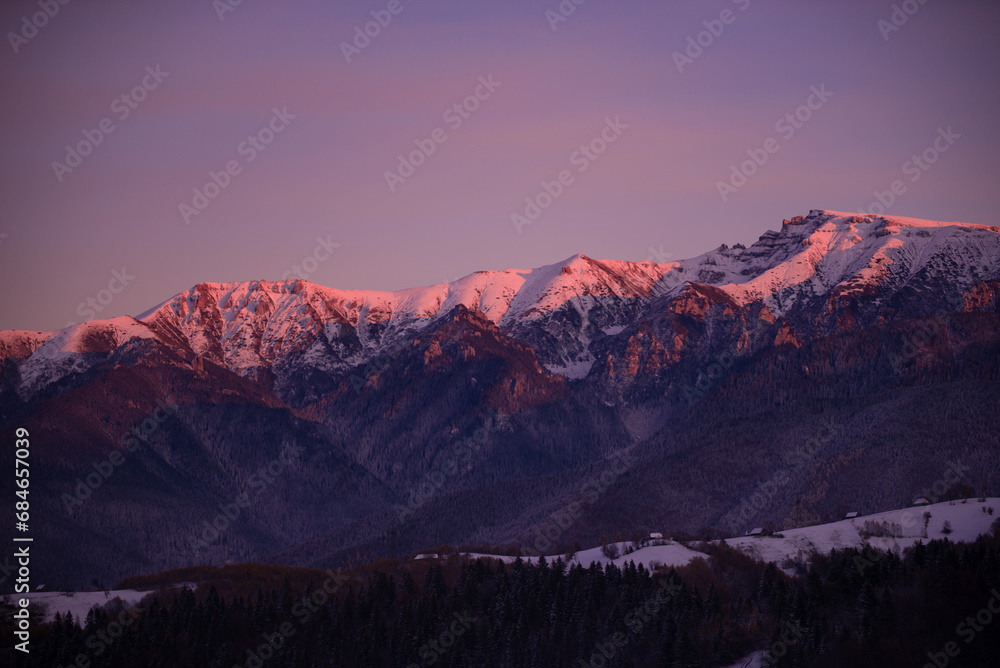 Fairy tale landscape with a rocky mountain full of snow at sunset. A village of traditional Romanian old houses spread over a valley.