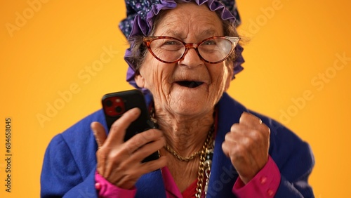 Closeup of toothless elderly senior old woman with wrinkled skin looking at phone having great happy success winner isolated on yellow background. People emotions, lifestyle concept. photo