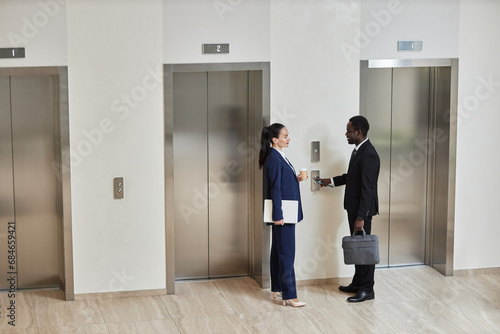 Wide angle view at Caucasian businesswoman with cup and laptop talking to cheerful African American businessman with phone and briefcase dressed in suits standing by elevators in lobby, copy space
