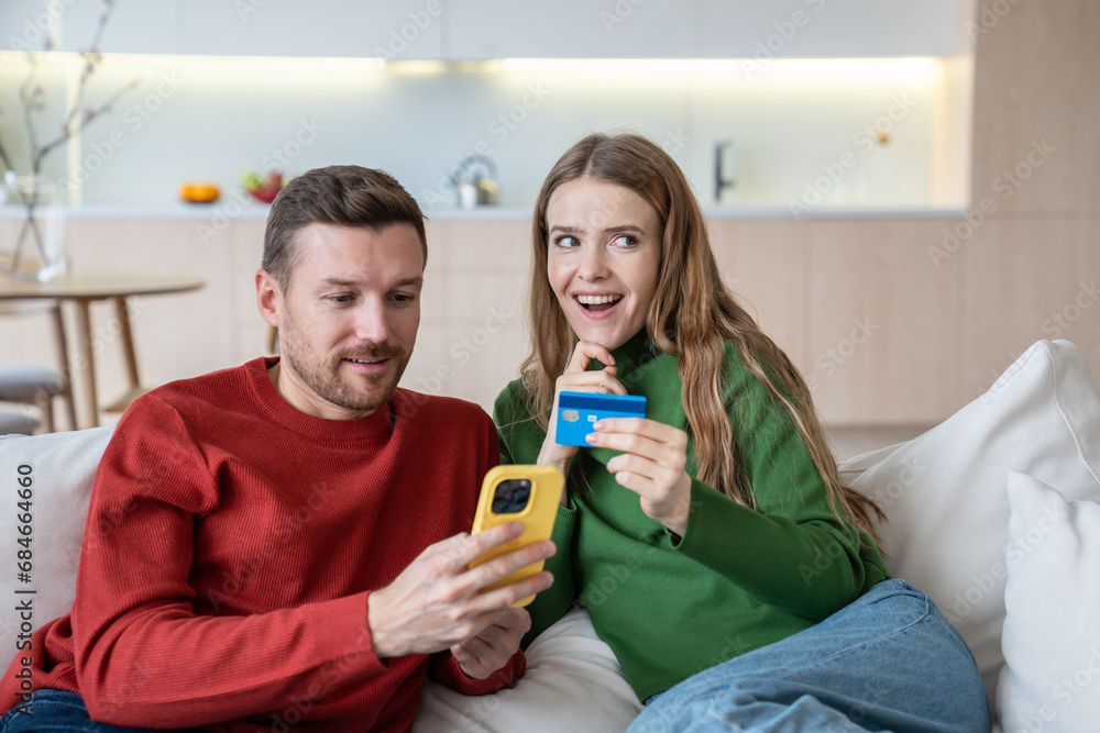 Happy man woman doing online payment in smartphone sitting on couch at home. Woman holds bank card with excited expecting face dreaming about purchase. Internet shopping buying choosing goods concept. Happy man woman doing online payment in smartphone sitting on couch at home. Woman holds bank card with excited expecting face dreaming about purchase. Internet shopping buying choosing goods concept.