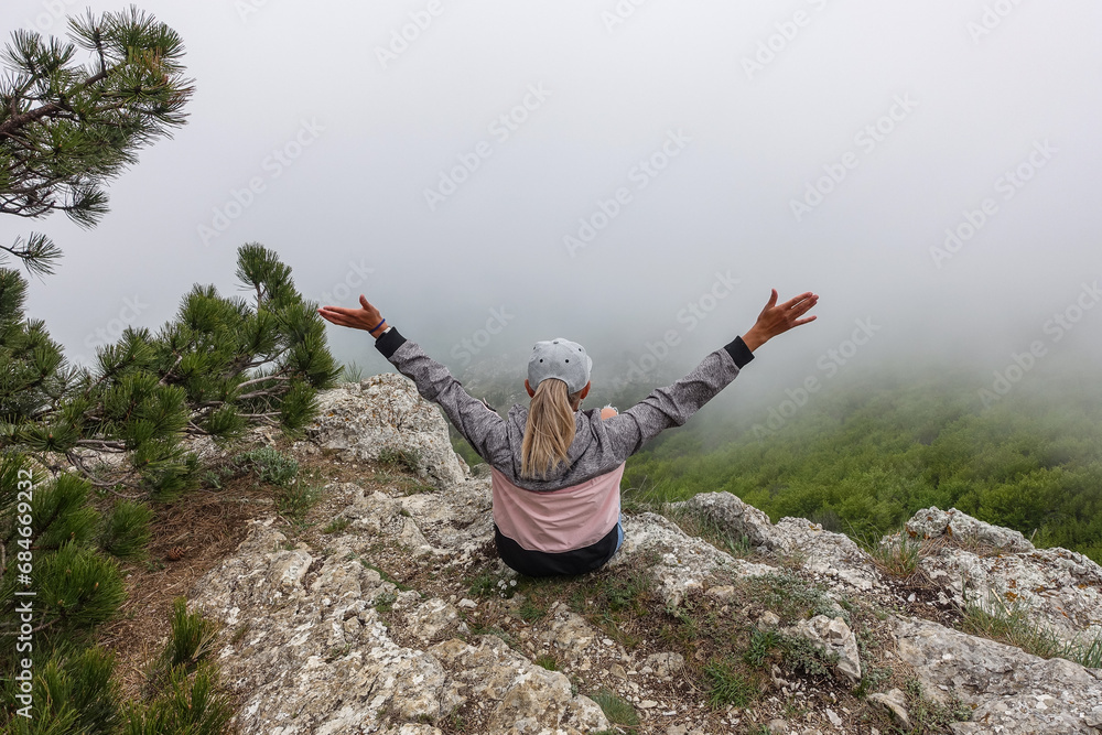 Obraz premium A picturesque mountain landscape in the clouds on Ai-Petri mountain in the Crimea. A woman on the background of a high-altitude landscape with trees in the clouds. Fog on the mountain.