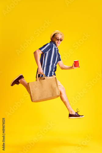 Happy young man, tourist in striped shirt, shirts, straw hat and sunglasses jumping, drinking coke against yellow studio background. Concept of human emotions, youth, summer vacation, fashion
