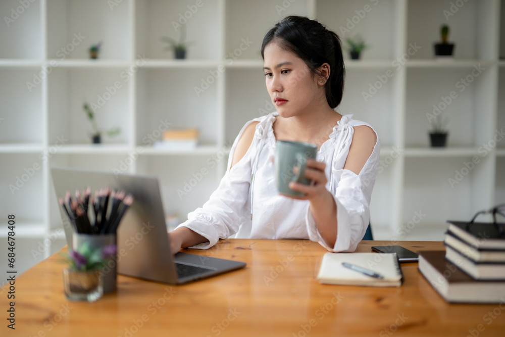 Asian woman sitting absent-minded pondering and looking laptop screen while taking a break from the job with coffee. feeling bored and tired Asian woman sitting absent-minded pondering and looking laptop screen while taking a break from the job with coffee. feeling bored and tired