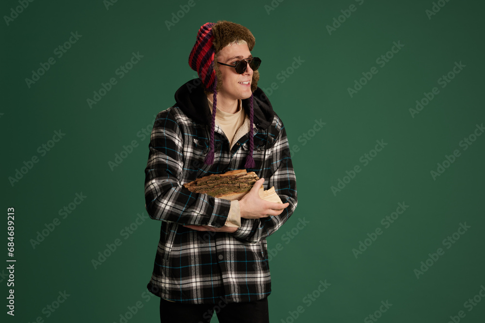 Smiling young man in checkered jacket, warm hat and sunglasses holding firewood against green studio background. Concept of human emotions, youth, winter fashion, vacations holidays Smiling young man in checkered jacket, warm hat and sunglasses holding firewood against green studio background. Concept of human emotions, youth, winter fashion, vacations holidays