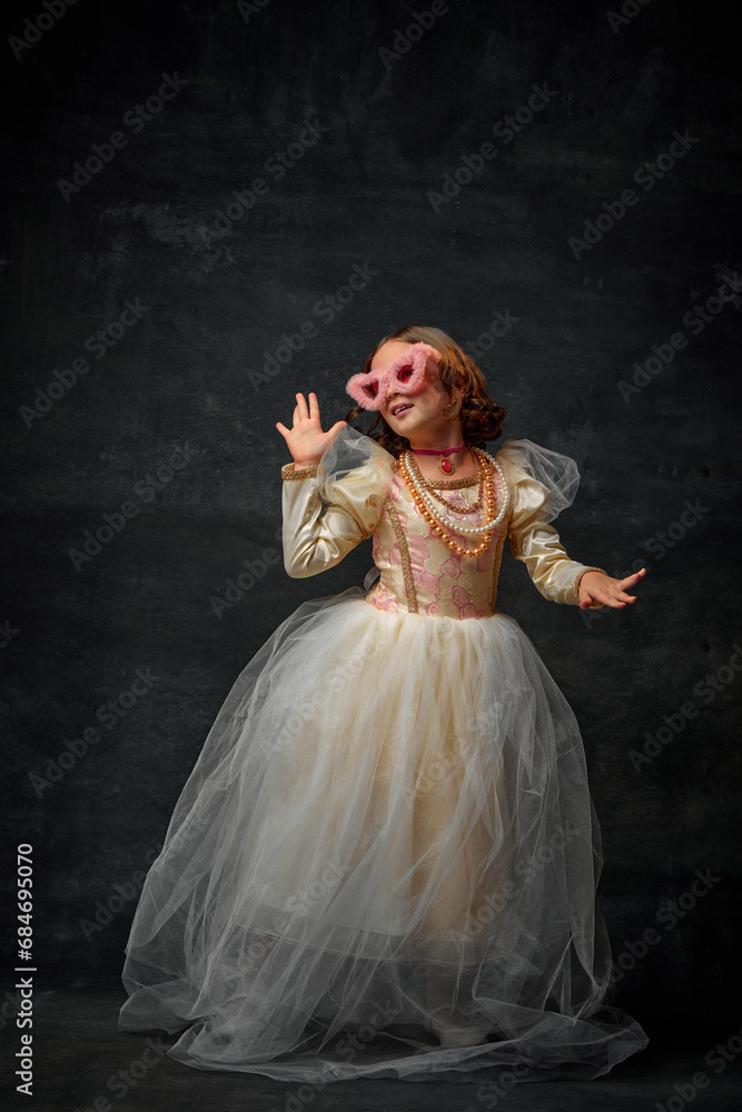 Full-length portrait of royal person, small girl dressed in old-fashioned costume with modern accessories an dancing against vintage background. Full-length portrait of royal person, small girl dressed in old-fashioned costume with modern accessories an dancing against vintage background.