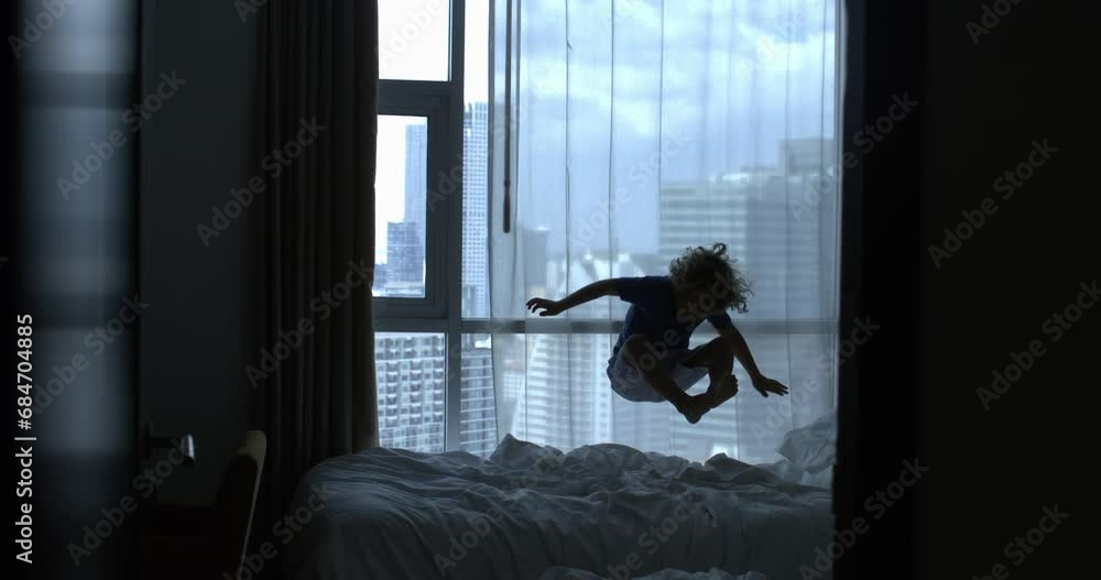 Super slow motion of carefree little boy is having fun to jump on bed at home in complete freedom and happiness in hotel room with view of skyscrapers during holidays family vacation trip. Super slow motion of carefree little boy is having fun to jump on bed at home in complete freedom and happiness in hotel room with view of skyscrapers during holidays family vacation trip.