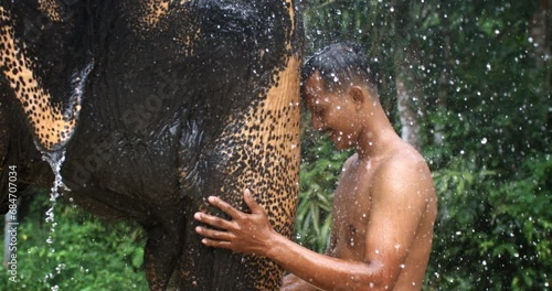 Super slow motion close up of young man taking care of wild rescued indian elephant under falling rain water drops on it's natural habitat tropical rainforest nature background during raining season. photo