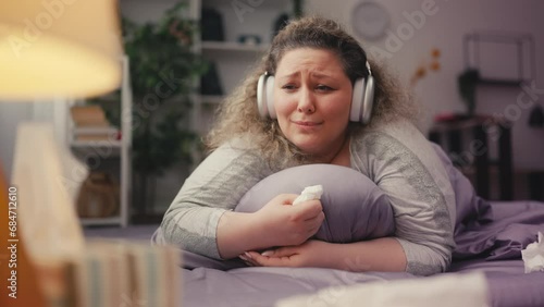 Sad plus-size woman crying on bed, listening to sentimental music in earphones photo
