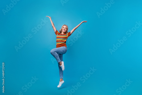 Full length photo of excited cheerful girl dressed print shirt jumping high rising arms empty space isolated blue color background