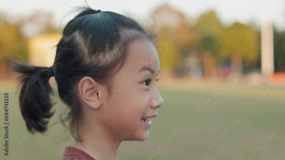Closed up face with Slow motion and moving of adorable 7-year-old Asian girl cycles bicycle through the lush greenery of a greenery grass playground, showing healthy and joyous childhood. Closed up face with Slow motion and moving of adorable 7-year-old Asian girl cycles bicycle through the lush greenery of a greenery grass playground, showing healthy and joyous childhood.