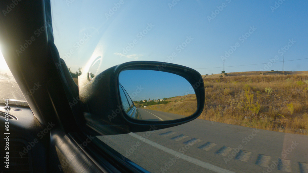 View from the car in the side mirror. A car is driving on a tarmac road in the countryside. There are yellow fields on the side of the road. The mirror reflects the road and the blue sky.