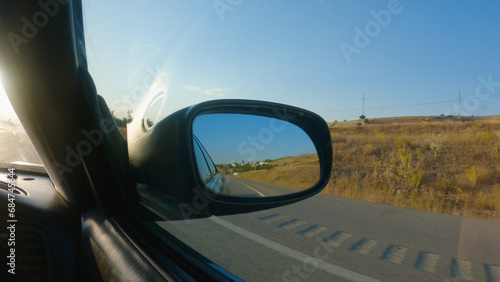 View from the car in the side mirror. A car is driving on a tarmac road in the countryside. There are yellow fields on the side of the road. The mirror reflects the road and the blue sky.