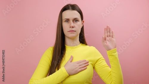 Serious conscious young woman 25s holding hand on chest and raising another swearing, trust and honest, patriotism, wearing yellow sweater, posing isolated over pink color background wall in studio