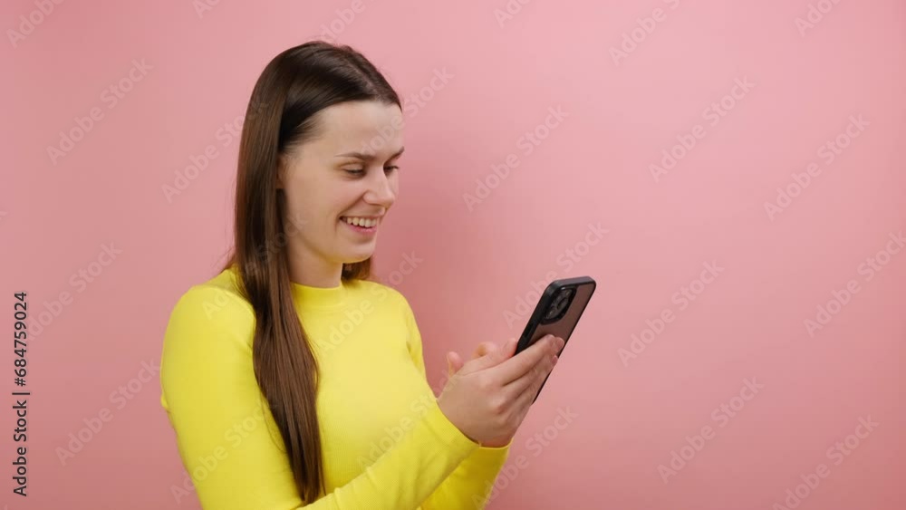 Smiling pretty young woman appearance scrolling social network posts on smartphone, user satisfied with mobile phone, wearing yellow sweater, posing isolated over pink color background wall in studio Smiling pretty young woman appearance scrolling social network posts on smartphone, user satisfied with mobile phone, wearing yellow sweater, posing isolated over pink color background wall in studio