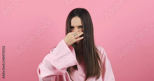 Disgusting odor. Discontent brunette woman frowning face, smells something awful, feeling aversion, pink studio background. Girl face with aversion dislikes something unpleasant. Woman wear pink dress photo