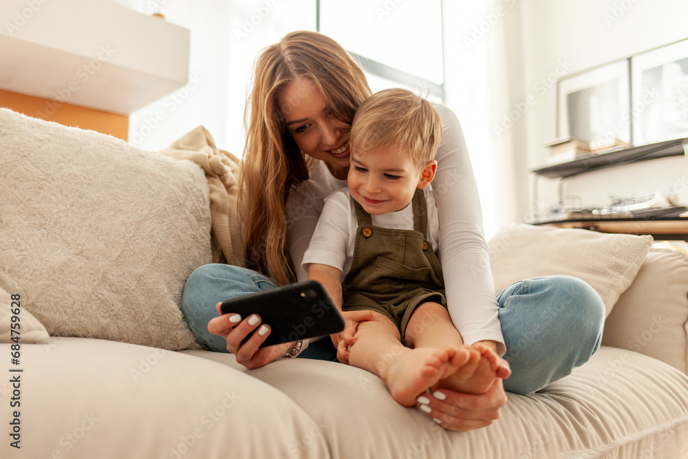 mother and her little son are sitting on the sofa and using smartphone, 2-year-old boy is looking at his mobile mother and her little son are sitting on the sofa and using smartphone, 2-year-old boy is looking at his mobile
