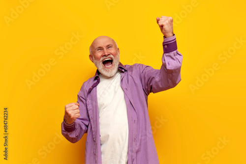 old bald grandfather in purple shirt celebrates victory with his mouth open on yellow isolated background photo