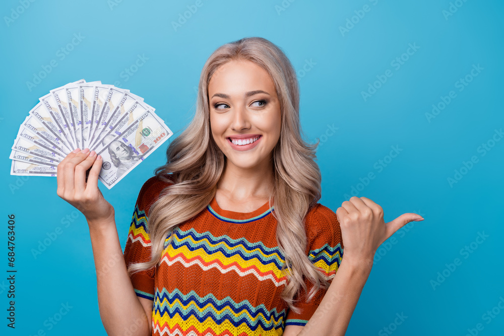 Photo of pretty dreamy girl dressed print shirt holding money fan looking showing thumb empty space isolated blue color background Photo of pretty dreamy girl dressed print shirt holding money fan looking showing thumb empty space isolated blue color background