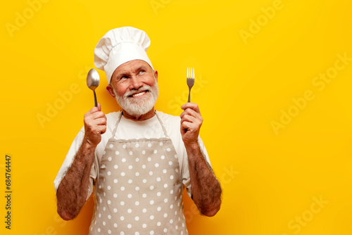 pensive old grandfather chef in hat and apron holds fork and spoon and dreams on yellow isolated background photo