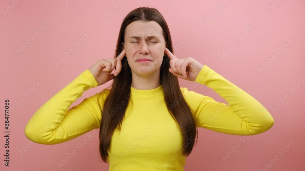 Nervous young caucasian woman standing and putting finger on ears and don't want to hear, nods her head, saying no, wearing yellow sweater, posing isolated over pink color background wall in studio Nervous young caucasian woman standing and putting finger on ears and don't want to hear, nods her head, saying no, wearing yellow sweater, posing isolated over pink color background wall in studio
