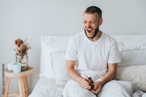 Sleepy young caucasian man in bed yawns holds jar with seeping pills, feels fatigue, needs healthy sleep. Pharmacology, medicine, recovery. Mockup, mental balance. photo