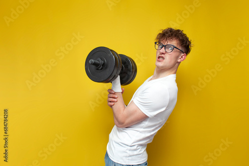 young nerd guy in glasses holds heavy dumbbells and strains on a yellow background, a motivated guy goes in for sports