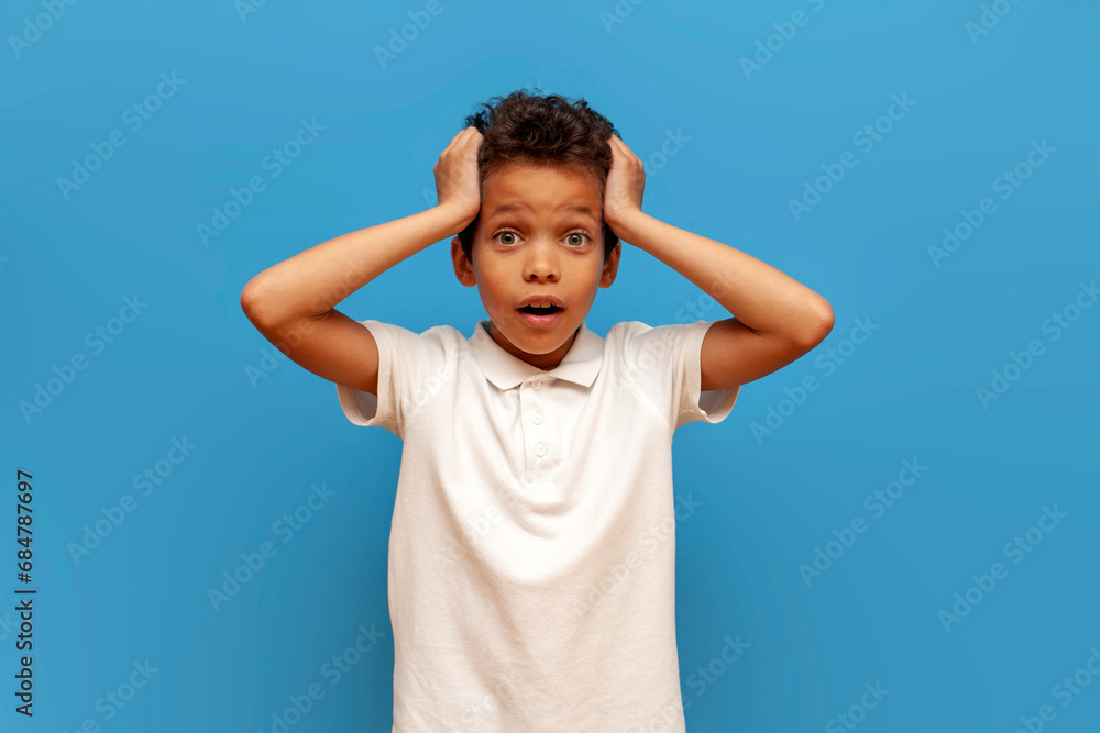 african american shocked teenage boy in white polo holding his head with his hands and looking at the camera african american shocked teenage boy in white polo holding his head with his hands and looking at the camera