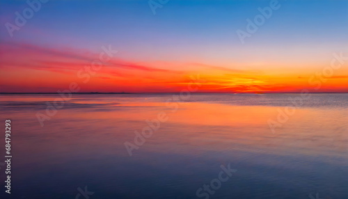 Gradient of colors in the sky as the sun sets, transitioning from warm oranges and pinks near the horizon to cool blues and purples higher in the sky. Emphasize the peaceful expanse of water. © emaotx
