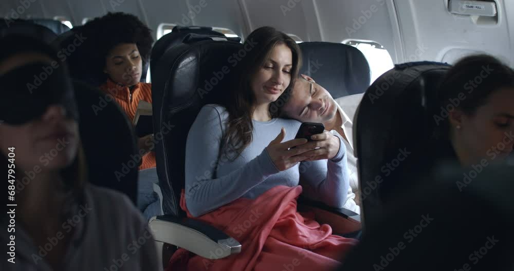 cute couple of friends in airplane have a nice moment together watching pictures, video call and smiling with sleepy boyfriend during the flight from the distant airport. cute couple of friends in airplane have a nice moment together watching pictures, video call and smiling with sleepy boyfriend during the flight from the distant airport.