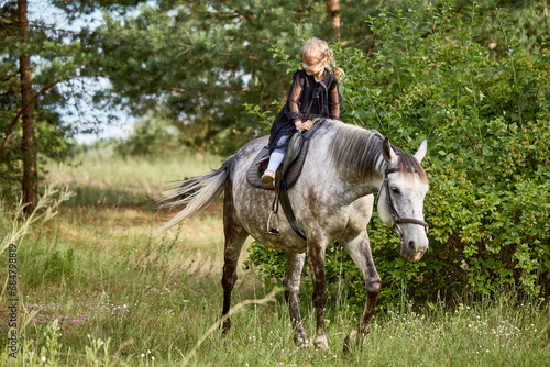 A little girl rides a horse outdoors. Cute girl riding a pony on a ranch. A child trains to ride a pet pony in the field.