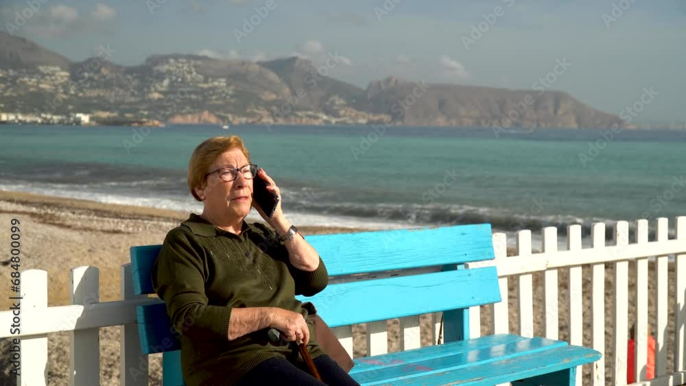 Old woman talking relaxed on phone, sitting on a bench near the beach, on a sunny morning. Old woman talking relaxed on phone, sitting on a bench near the beach, on a sunny morning.