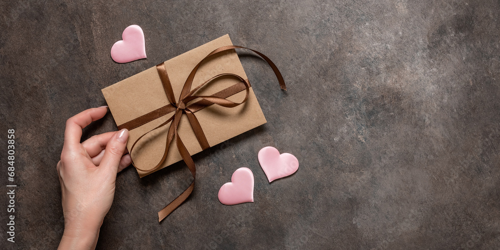 Female hand holds a brown envelope tied with a silk ribbon and pink silk hearts on a dark vintage background. Valentine's Day card. Top view, flat lay, copy space. Banner - obrazy, fototapety, plakaty 