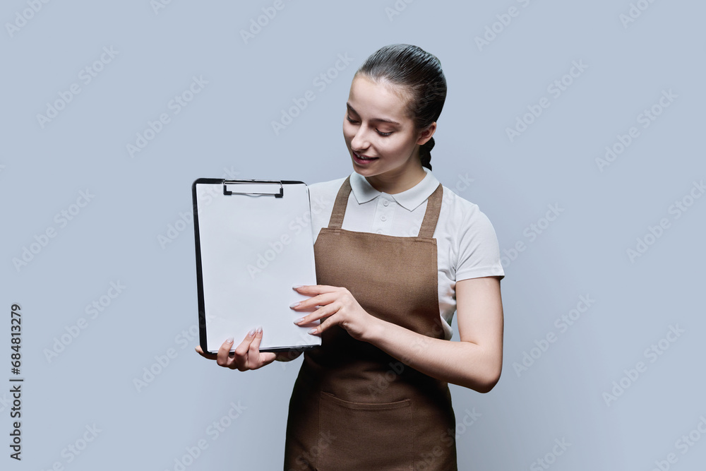 Young female worker in apron with blank sheet on clipboard, on gray background Young female worker in apron with blank sheet on clipboard, on gray background