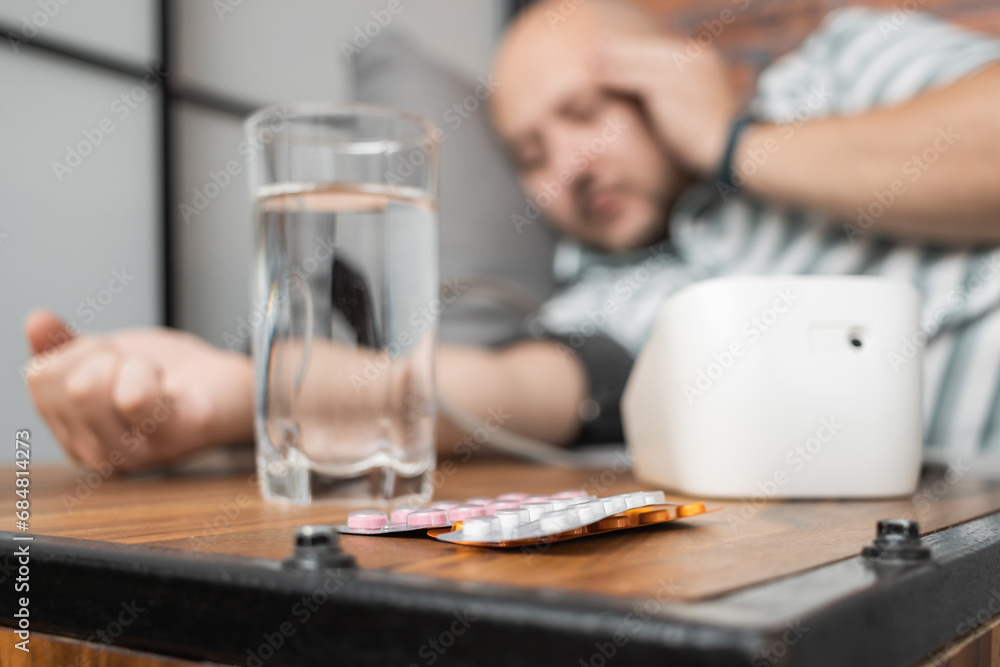 Defocused background with man who has hypertension and headache, lying on sofa at home, measures blood pressure on hand with modern digital tonometer, focus on blister pack with pills on foreground Defocused background with man who has hypertension and headache, lying on sofa at home, measures blood pressure on hand with modern digital tonometer, focus on blister pack with pills on foreground
