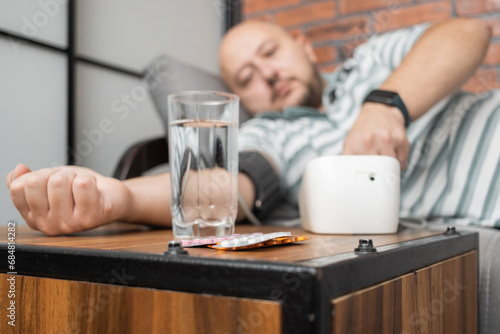 Defocused background with man who has hypertension and headache, lying on sofa at home, measures blood pressure on hand with modern digital tonometer, focus on blister pack with pills on foreground