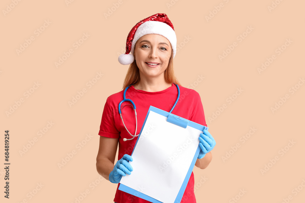 Mature female doctor in Santa hat with clipboard on beige background Mature female doctor in Santa hat with clipboard on beige background
