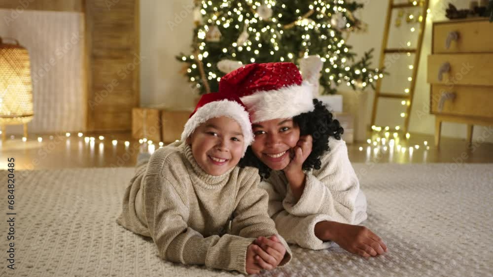 Smiling curly mother and kid in santa hats lying on stomachs in beautifully decorated living room with garlands, Christmas tree, warm yelow lights. High quality 4k footage Smiling curly mother and kid in santa hats lying on stomachs in beautifully decorated living room with garlands, Christmas tree, warm yelow lights. High quality 4k footage