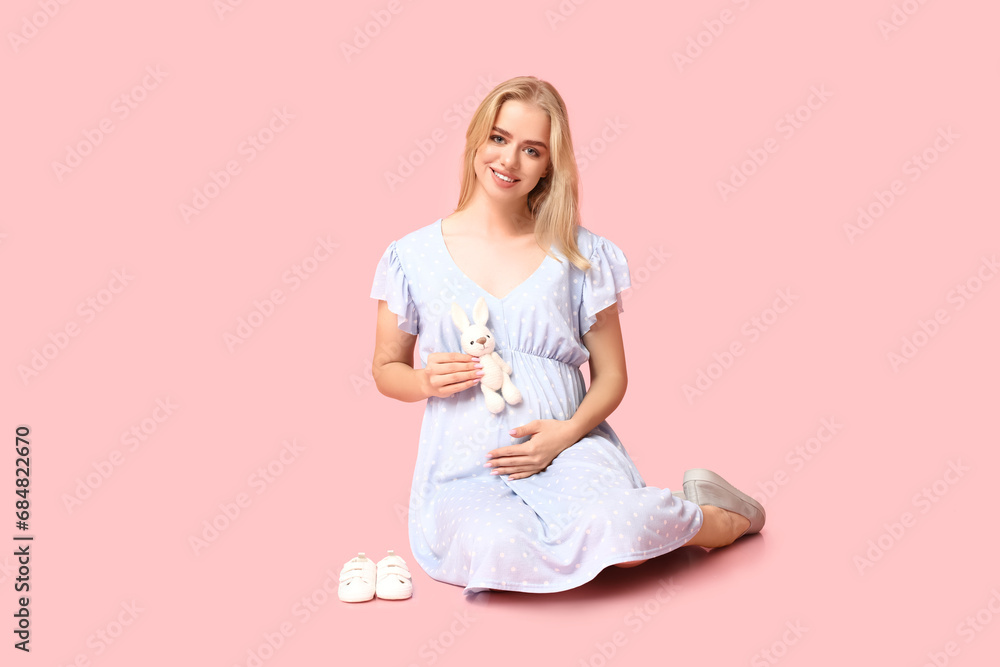 Beautiful young pregnant woman with baby shoes and toy sitting on pink background Beautiful young pregnant woman with baby shoes and toy sitting on pink background