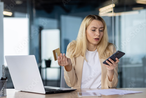 Frustrated sad upset woman working inside office, cheated business woman refused online money transfer, female worker displeased holding bank credit card and phone photo