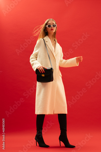 Fashionable confident woman wearing trendy white boucle midi coat, stylish sunglasses, over the knee high heel boots, carrying classic black leather shoulder bag, posing on red background