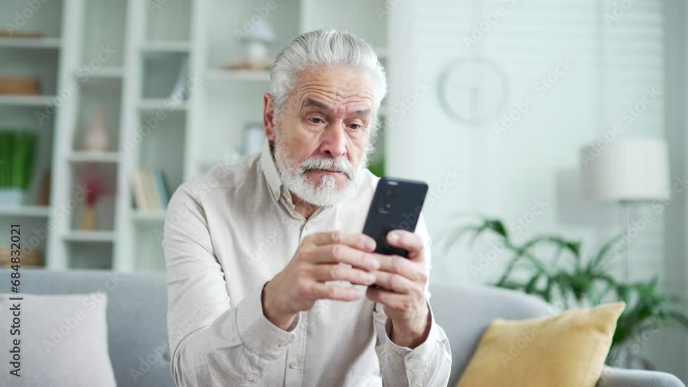 Frustrated stressed elderly senior gray haired man reads bad news on a smartphone sitting on sofa in living room at home. Worried depressed adult mature male is sad reviewing negative message on phone Frustrated stressed elderly senior gray haired man reads bad news on a smartphone sitting on sofa in living room at home. Worried depressed adult mature male is sad reviewing negative message on phone