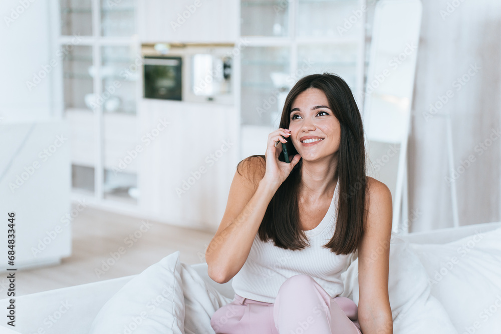 beautiful fashion model with Asian roots is sitting on couch at home talking on phone looking away smiling enjoying communication with a friend. A young American woman spends time at home on weekends beautiful fashion model with Asian roots is sitting on couch at home talking on phone looking away smiling enjoying communication with a friend. A young American woman spends time at home on weekends