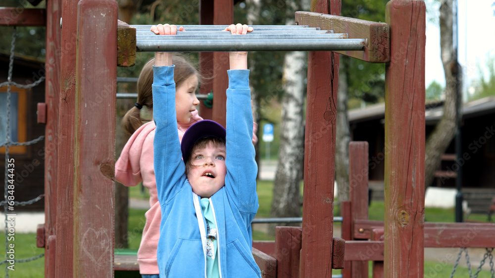 Fototapeta premium School age children two girls playing around on the playground, climbing, kids and fun physical exercise outdoors, sisters, siblings, hanging. Strength and agility, young age, real people lifestyle