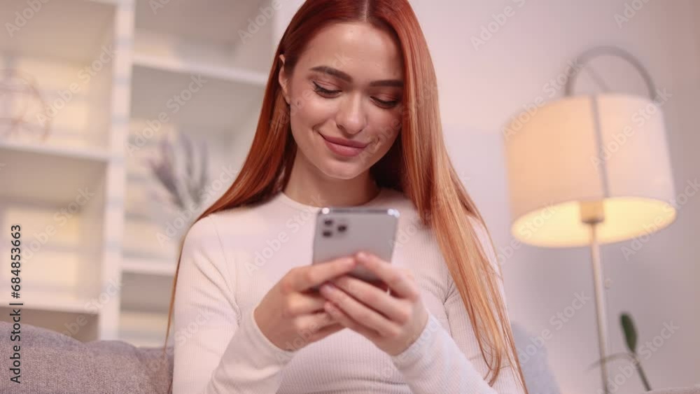Charming young red haired woman hold smartphone scrolling app watching social media feed and looking at the screen indoors Happy female texting enjoying leisure time at cozy home Charming young red haired woman hold smartphone scrolling app watching social media feed and looking at the screen indoors Happy female texting enjoying leisure time at cozy home