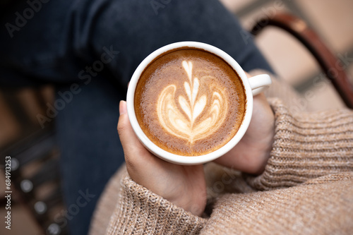 A woman in a cosy knitted sweater holding a cup of hot cappuccino, enjoying morning coffee at cafe.