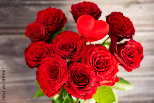 Close-up of a bouquet of red roses with green leaves and a decorative red heart against wooden background