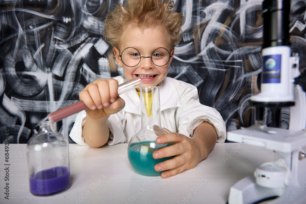 Cute blond boy participates in scientific study and conducts chemical scientific experiment by analyzing and mixing liquid in test tube. Boy mixes substances different colors in cones different shapes Cute blond boy participates in scientific study and conducts chemical scientific experiment by analyzing and mixing liquid in test tube. Boy mixes substances different colors in cones different shapes