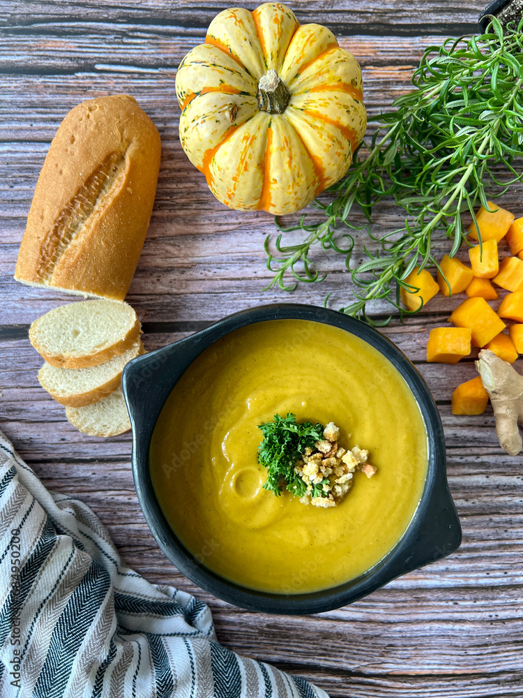 Flat lay of butternut winter squash soup on textured wooden surface with bread and rosemary suitable for Fall and winter background. 