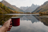 a man holds a red coffee mug against the background of an autumn forest and a mountain lake