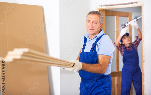 Middle-aged male carpenter in a blue uniform holding wooden planks indoors © JackF